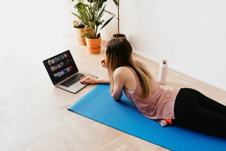young woman lying on floor on a pilates mat while using laptop at home